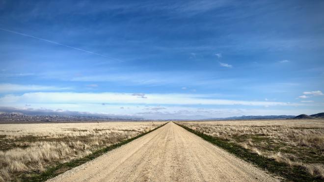 A landscape photograph with a dirt road in the foreground heading straight to the horizon with golden grass on either side. Bright blue sky above with wispy clouds and a contrail.